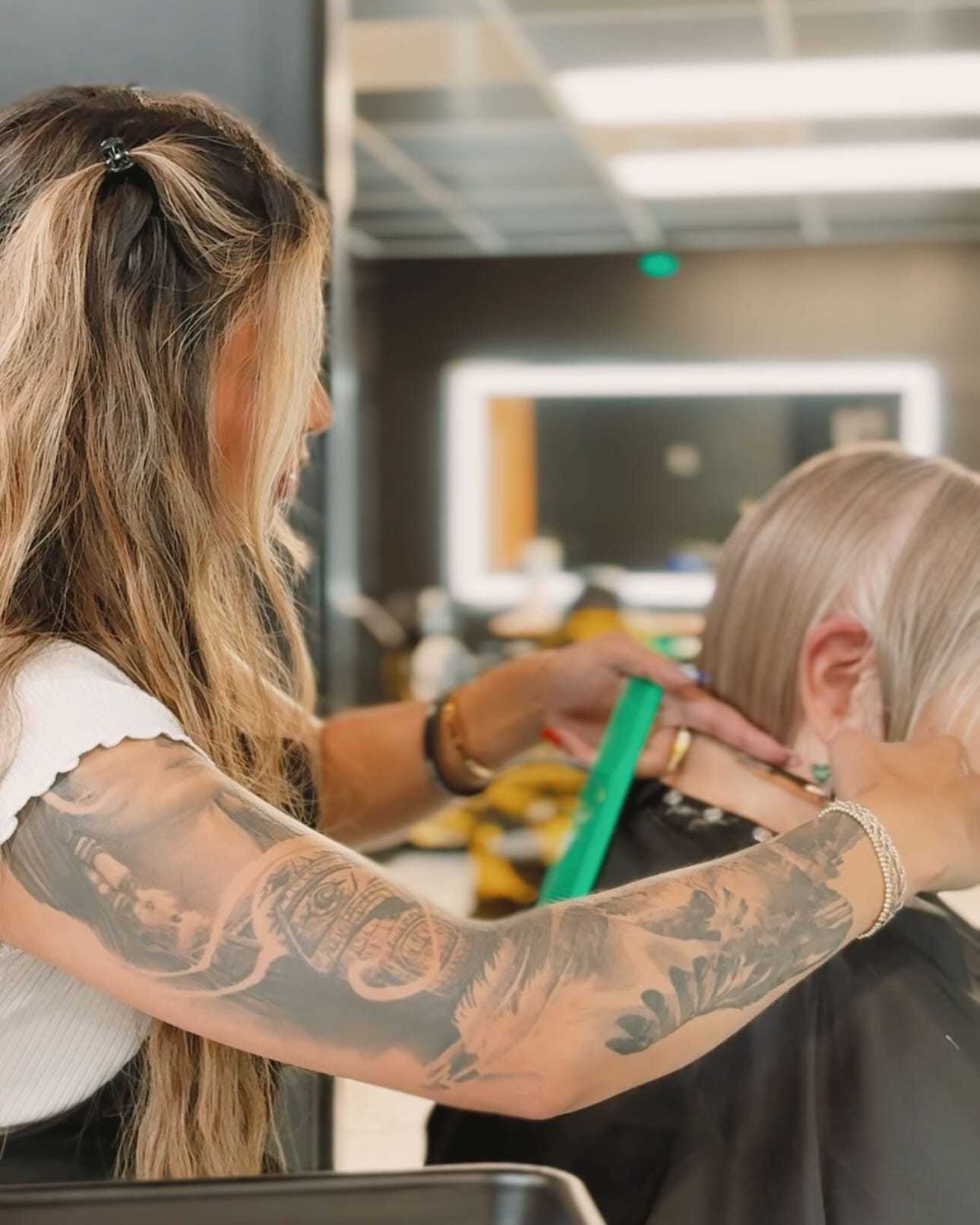 Hairstylist with tattoos trims client's short blonde hair in modern salon.